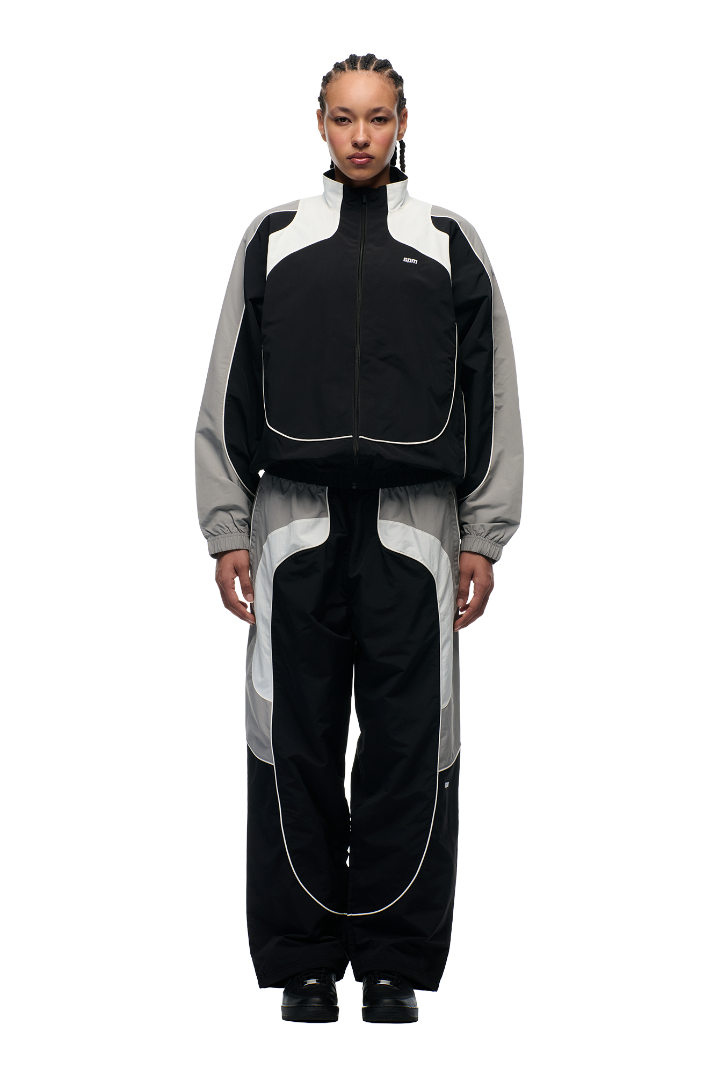 A person with braided hair faces forward, wearing the 6PM EVERYDAY TRACK JACKET BLACK featuring white piping, embroidered logos, mesh lining, and black shoes, set against a plain light background.