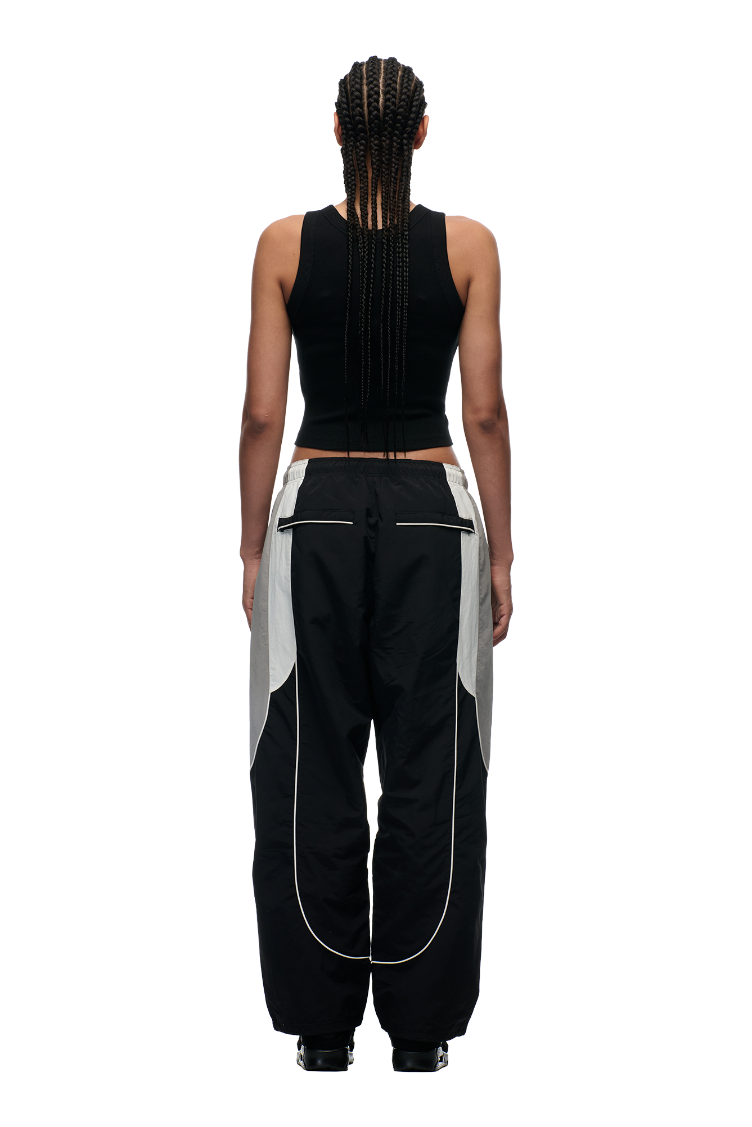 A person with long braids is shown from the back against a plain white background, wearing 6PM's EVERYDAY TRACK PANTS BLACK with gray and white panels, zipped pockets, and a black sleeveless crop top.