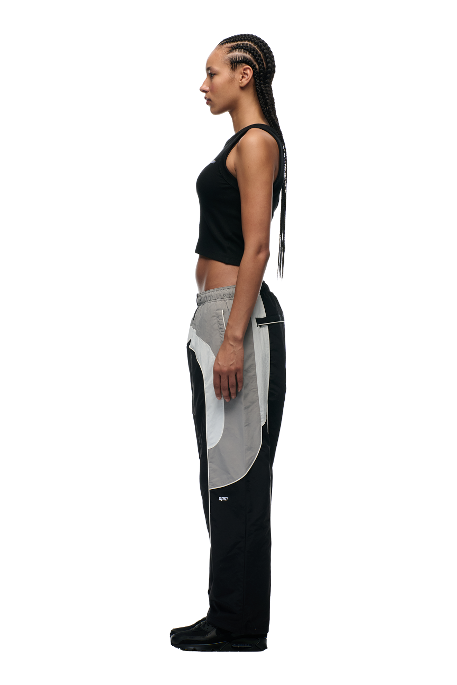 A woman with long braids stands in profile wearing 6PM’s EVERYDAY TRACK PANTS BLACK, featuring zipped pockets and a color-blocked design, paired with a black sleeveless crop top and black shoes against a plain white background.