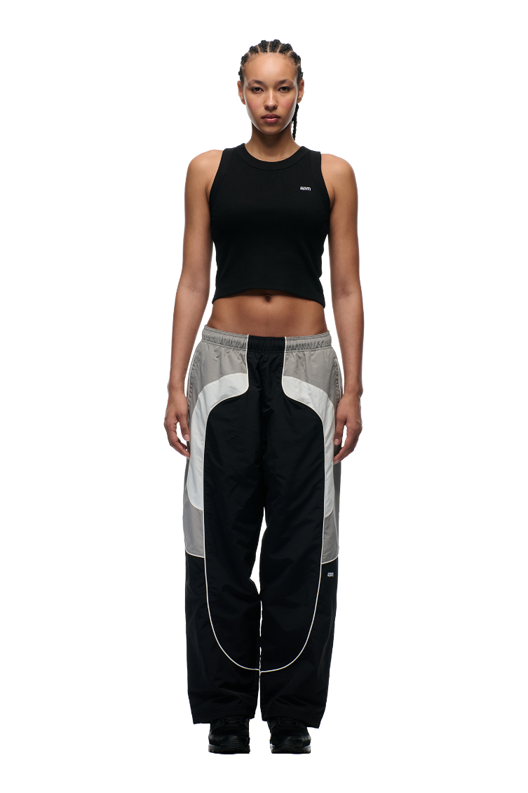 A person with braided hair wears the 6PM EVERYDAY TRACK PANTS BLACK, featuring zipped pockets and white piping, styled with a black sleeveless crop top while standing against a plain white background.
