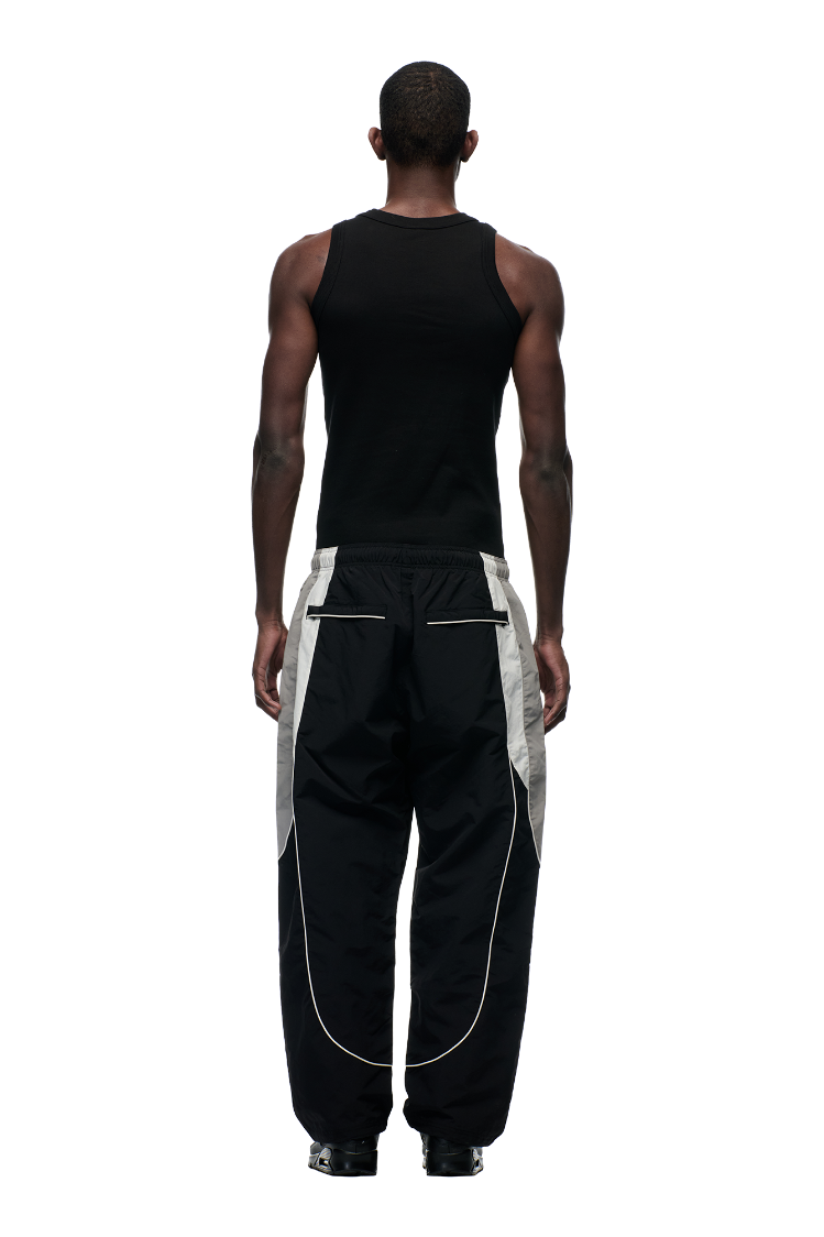 A person faces away, wearing 6PM's EVERYDAY TRACK PANTS BLACK—loose black pants with light gray side stripes, zipped pockets, and white piping—paired with a black sleeveless shirt against a plain white background.