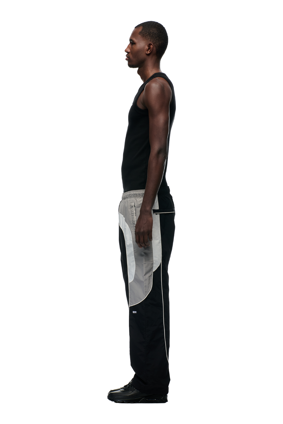 A man stands in profile against a plain background, wearing a black tank top and 6PM’s EVERYDAY TRACK PANTS BLACK—color-blocked with gray, featuring zipped pockets for convenience—and black shoes.