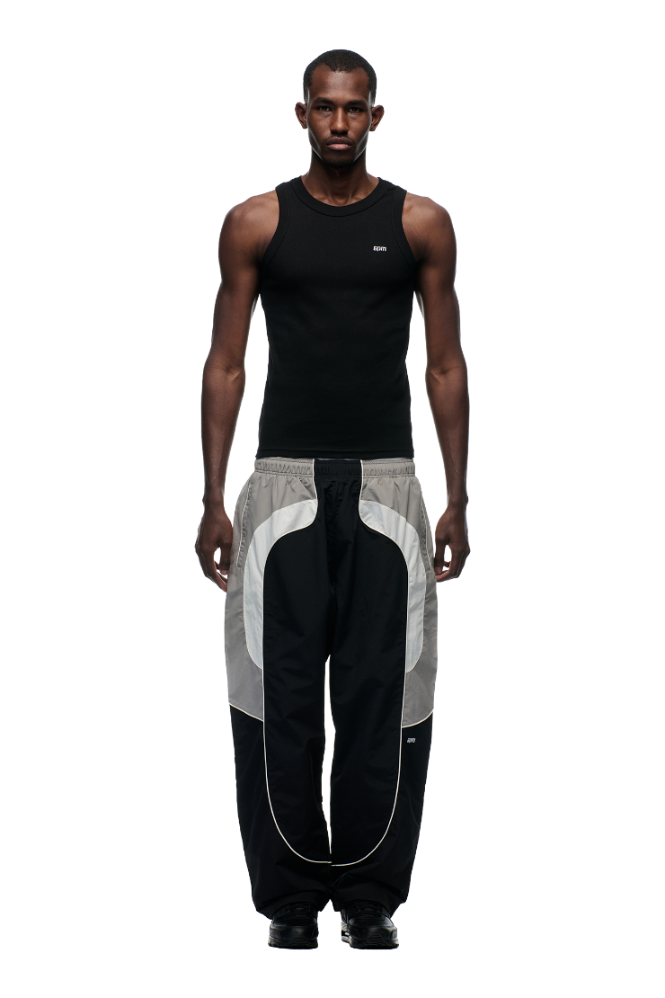 A man stands facing forward against a plain white background, wearing the 6PM EVERYDAY TRACK PANTS BLACK, featuring loose fit, white trim, and zipped pockets, paired with a black sleeveless tank top.