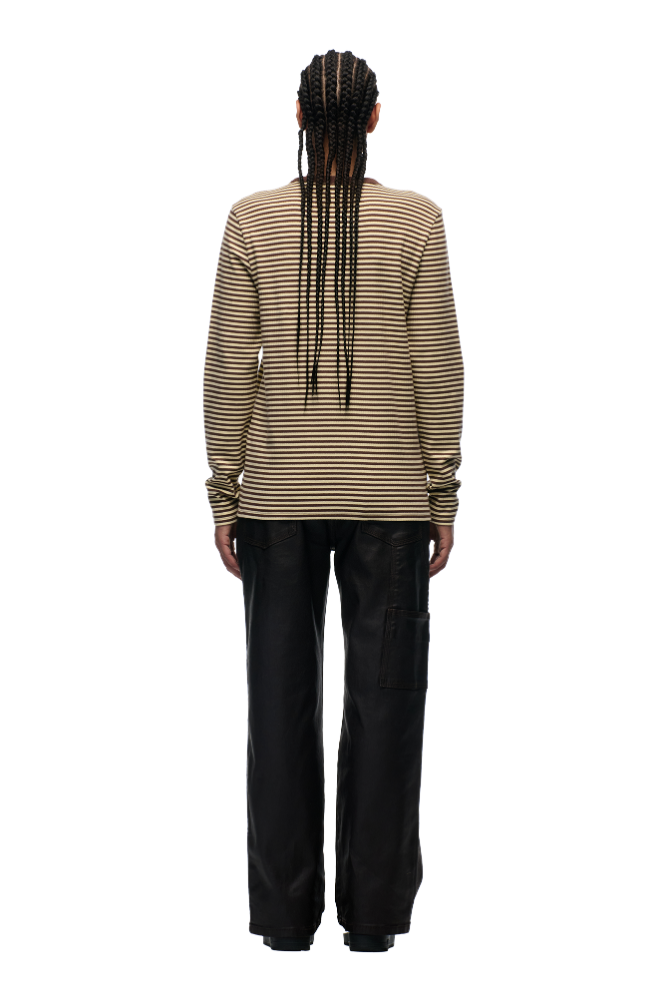 A person stands with their back to the camera, wearing 6PM’s STRIPED RIBBED LS DARK HAZELNUT top and loose black pants. Long braided hair complements the look, while a subtle tonal flag label accents the shirt against a plain white background.