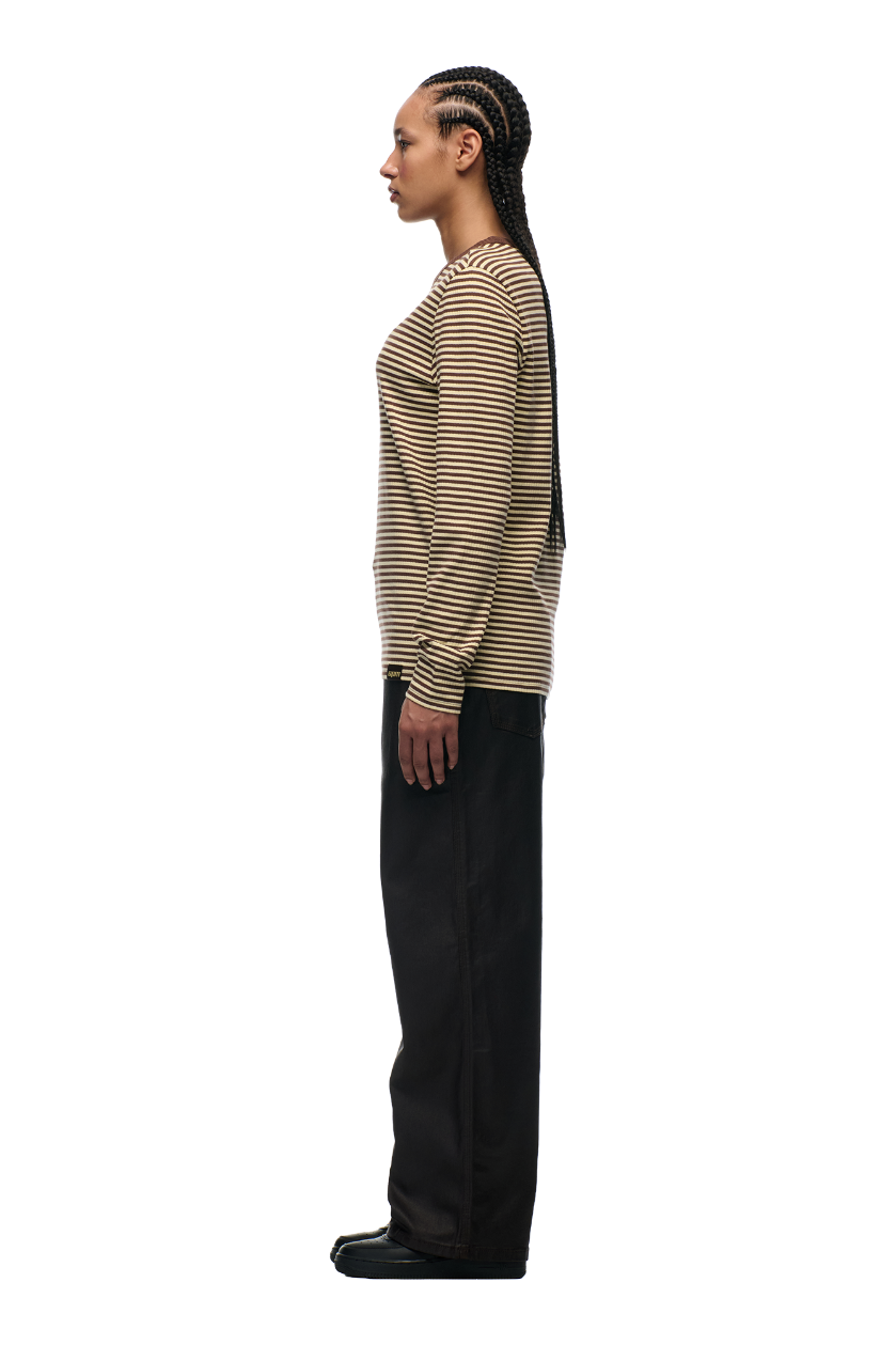 A person with long braids stands in profile against a plain background, wearing the 6PM STRIPED RIBBED LS DARK HAZELNUT top, loose black pants, and black shoes.