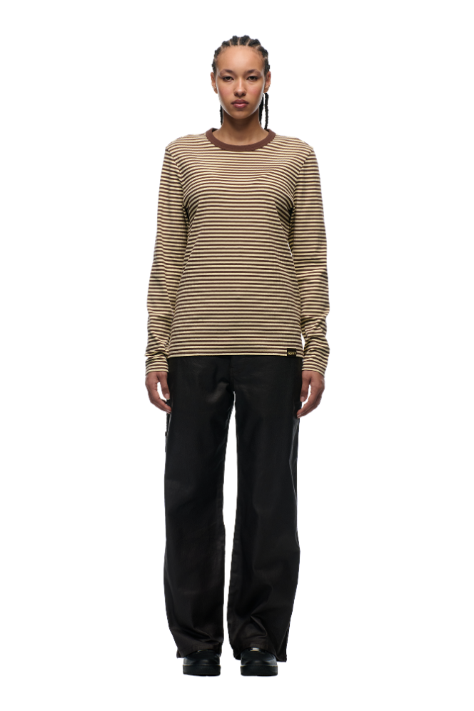 A person with braided hair stands facing forward in 6PM's STRIPED RIBBED LS DARK HAZELNUT top and loose black pants against a plain light background.