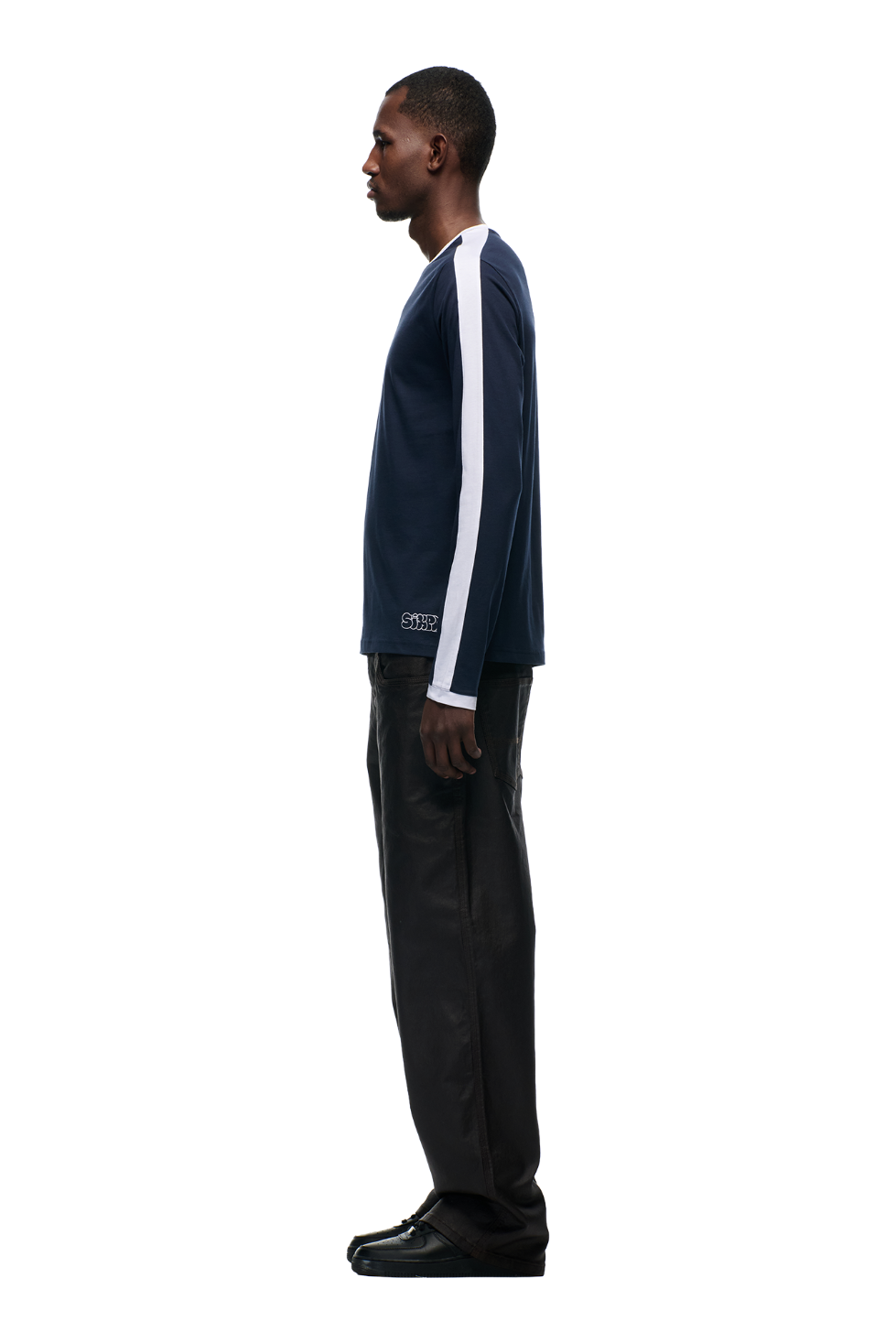 A man faces left in profile against a white background, wearing the 6PM GRAFFITI RINGER LS MIDNIGHT BLUE top with white arm stripes, paired with black pants and black shoes.