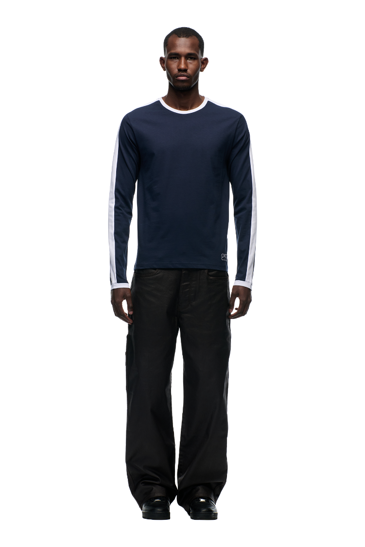 A man stands facing forward against a light gray background, wearing the 6PM GRAFFITI RINGER LS MIDNIGHT BLUE top, loose black pants, and black shoes.