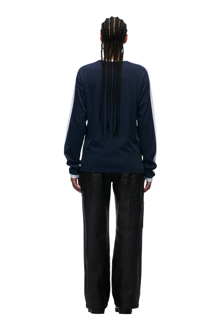 A person with long braids faces away, wearing the 6PM GRAFFITI RINGER LS MIDNIGHT BLUE top with color-block cuffs and white sleeve stripes, paired with loose black pants, against a plain light background.