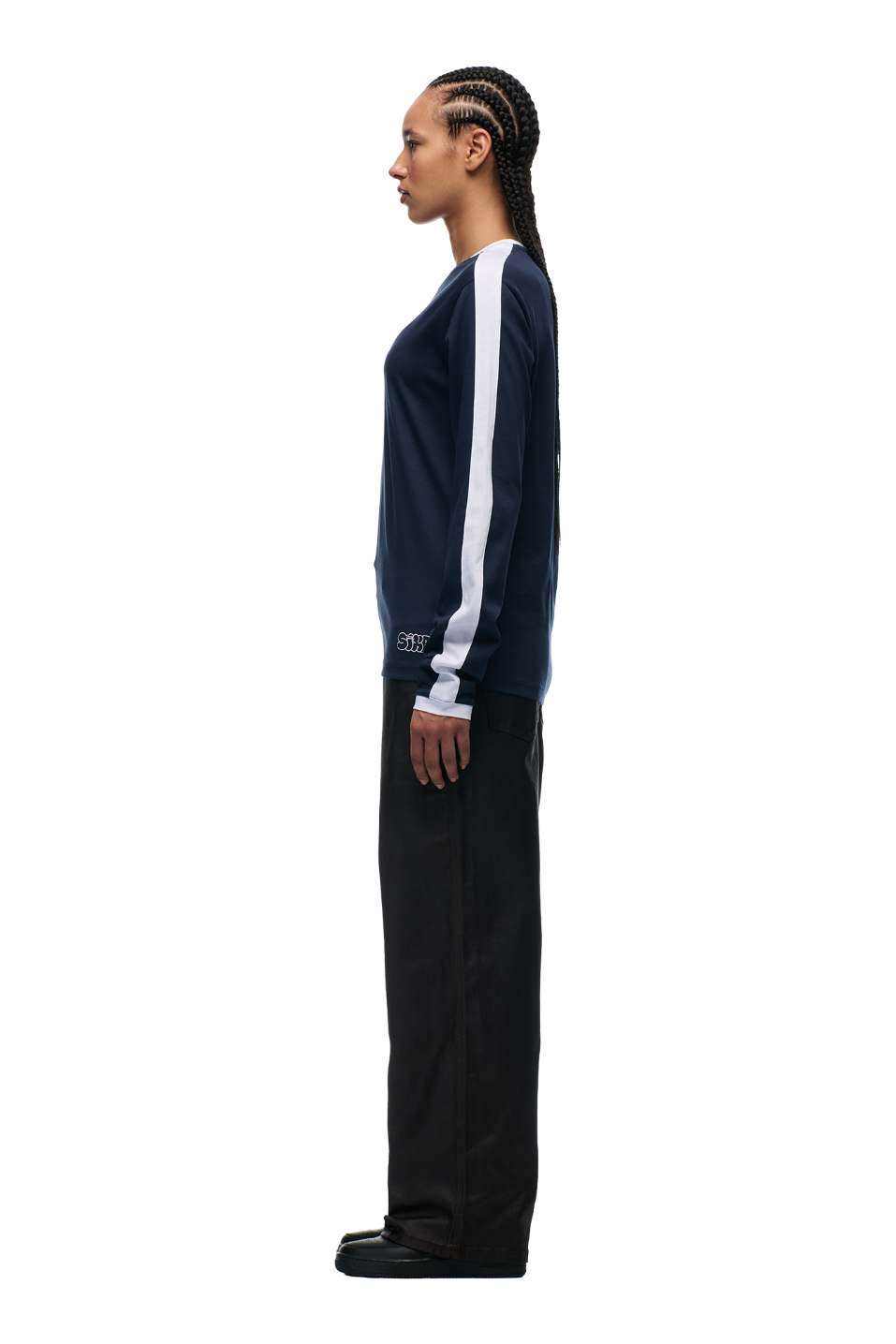 A person with braided hair stands in profile against a white background, wearing the 6PM GRAFFITI RINGER LS MIDNIGHT BLUE top, paired with black pants and black shoes.