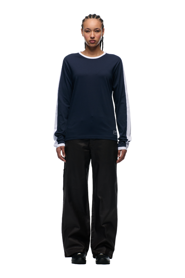 A person with braided hair wears the 6PM GRAFFITI RINGER LS MIDNIGHT BLUE, loose black pants, and black shoes, standing against a plain white background.
