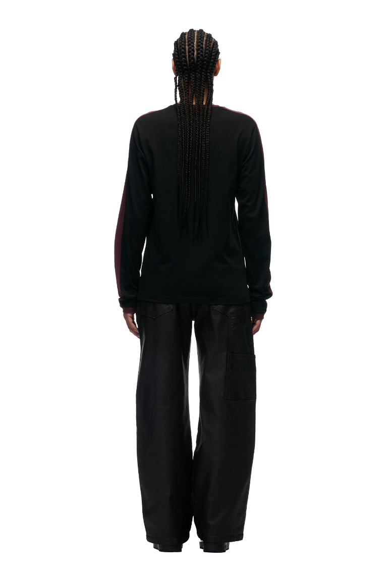A person with long, braided hair faces away, wearing the 6PM GRAFFITI RINGER LS BLACK shirt and loose-fitting black pants against a plain white background.