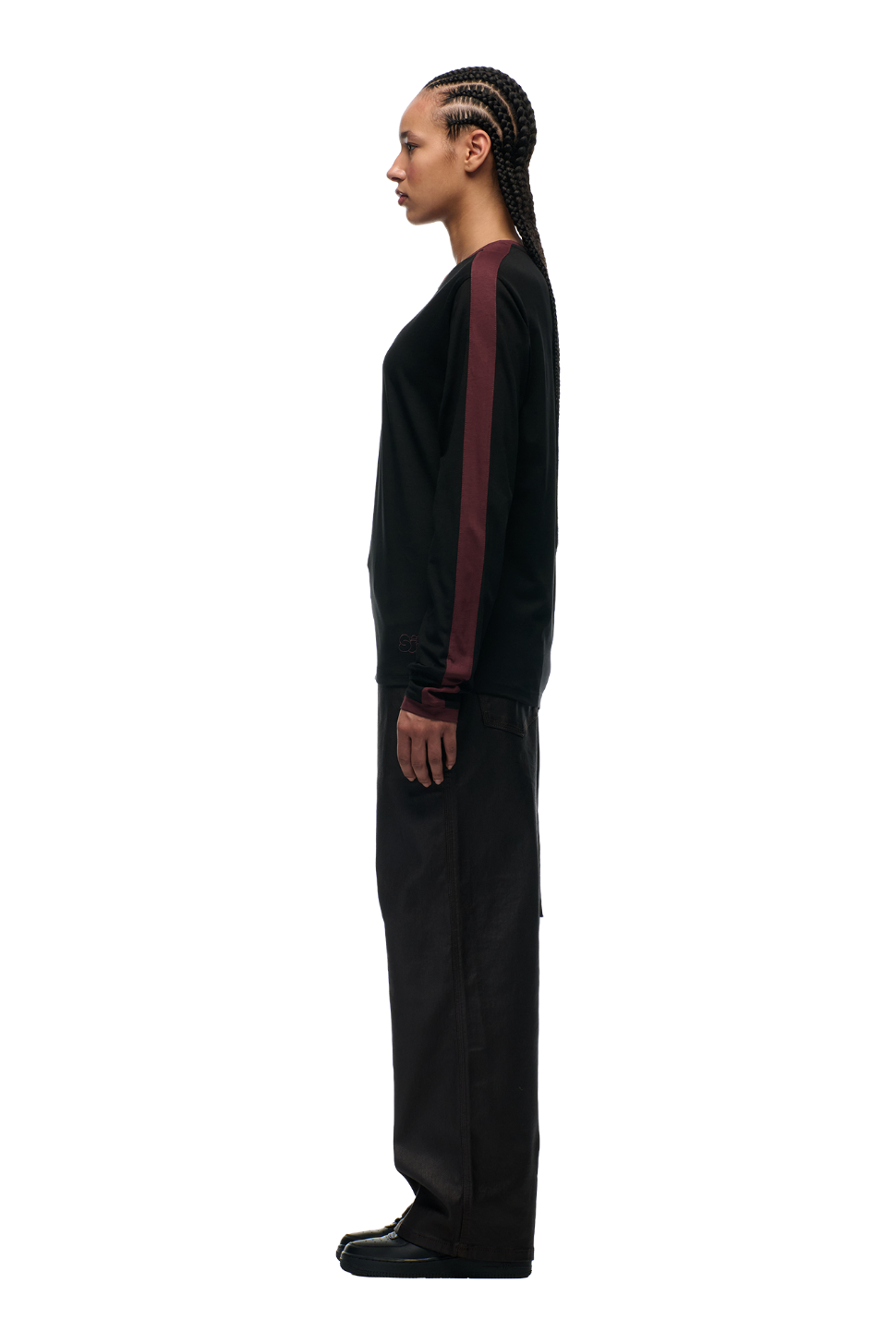 A person with braided hair stands in profile, wearing 6PM’s GRAFFITI RINGER LS BLACK top and loose black pants, set against a plain white background.