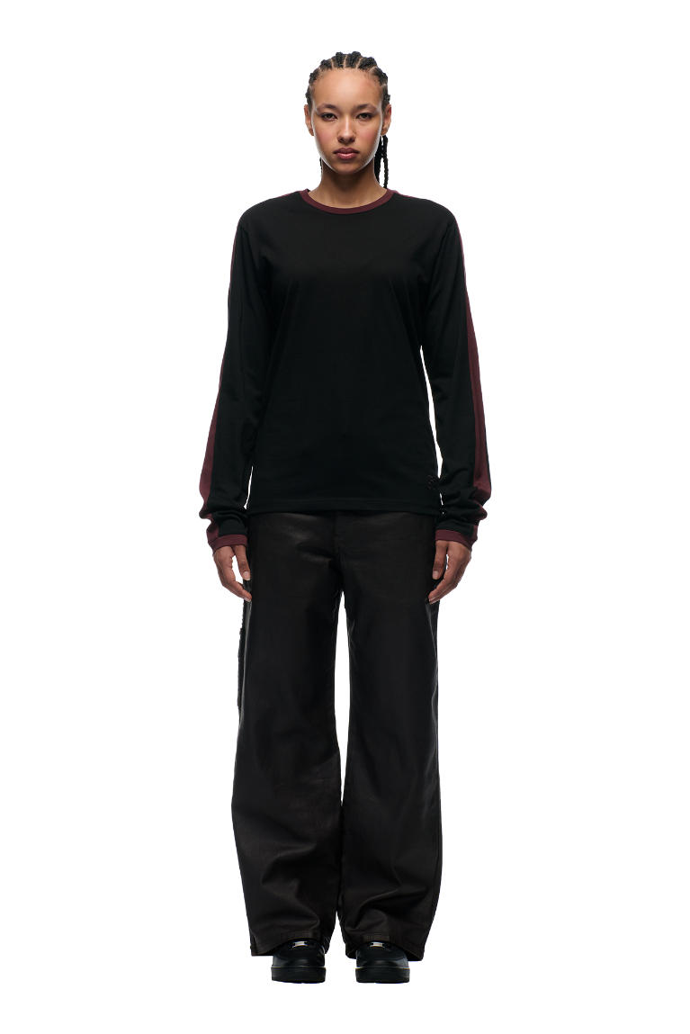 A person with braided hair faces forward, wearing the 6PM GRAFFITI RINGER LS BLACK long-sleeve shirt with color-block cuffs and maroon sleeves, paired with loose-fitting black pants, set against a plain light gray background.