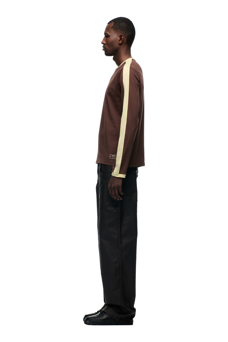A man stands in profile against a plain background, wearing the 6PM GRAFFITI RINGER LS DARK HAZELNUT top, featuring a brown body with light yellow sleeve stripes, paired with dark pants and black shoes.