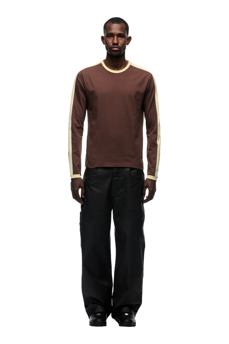 A man stands facing forward in the 6PM GRAFFITI RINGER LS DARK HAZELNUT long-sleeve shirt, paired with loose black pants and black shoes, against a plain white background.