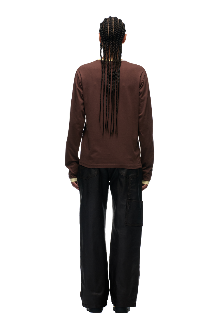 A person with long braids faces away, wearing the 6PM GRAFFITI RINGER LS DARK HAZELNUT long-sleeve shirt with color-block cuffs and loose black pants, set against a plain white background.