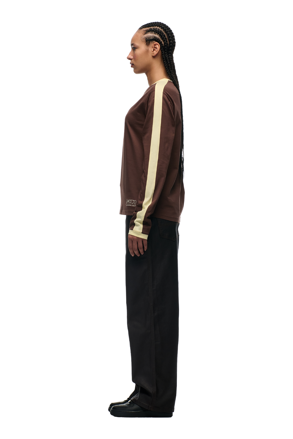 A person with long, braided hair stands in profile against a white background, wearing the 6PM GRAFFITI RINGER LS DARK HAZELNUT top, paired with black pants and black shoes.