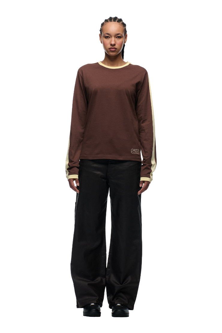 A person with braided hair wears the 6PM GRAFFITI RINGER LS DARK HAZELNUT long-sleeve top and loose black pants, standing facing forward against a plain white background.