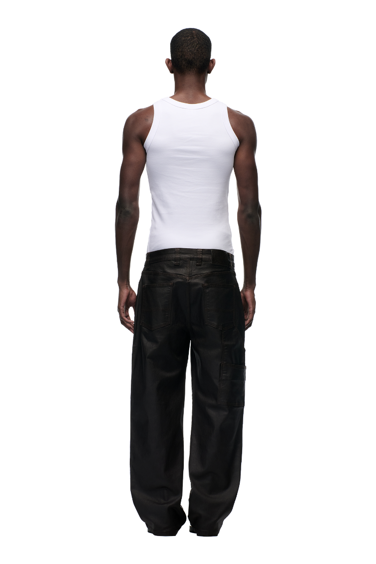 A man faces away from the camera in a fitted white tank top and 6PM's COATED CARPENTER DENIM HASH BROWNIE pants, set against a plain white background.