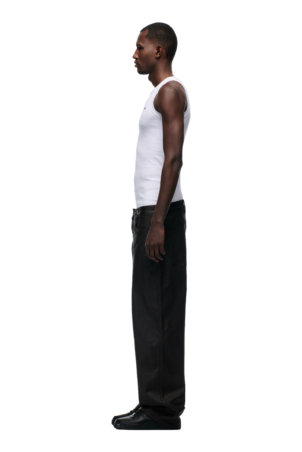 A man in profile faces left, wearing a fitted white tank, 6PM COATED CARPENTER DENIM HASH BROWNIE pants with metal buttons, and black shoes against a plain white background.