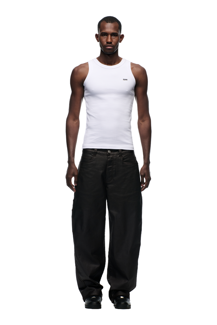 A man faces forward in a fitted white tank and 6PM’s COATED CARPENTER DENIM HASH BROWNIE pants, styled with black shoes, set against a plain white background.
