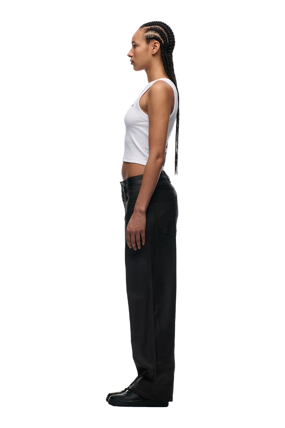 A woman with braided hair stands in profile, facing left, wearing a white sleeveless crop top and 6PM COATED CARPENTER DENIM HASH BROWNIE pants with metal buttons, plus black shoes, against a plain white background.