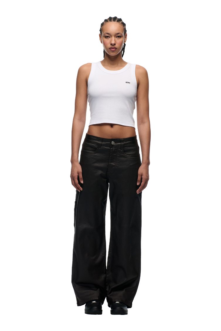 Facing forward, the person wears a white cropped tank with 6PM's COATED CARPENTER DENIM HASH BROWNIE pants. Black shoes finish the look, hair styled in tight braids, all against a plain white background.