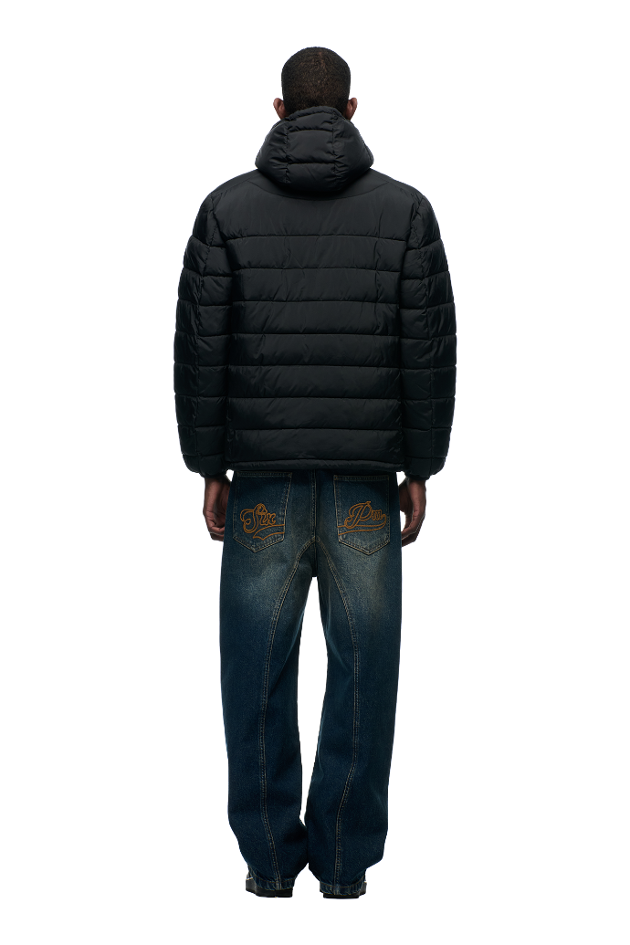 Facing away, a person wears the 6PM LIGHT WEIGHT PUFFER JACKET in black, featuring a hood, paired with loose blue jeans with yellow embroidery on the back pockets, set against a plain light background.