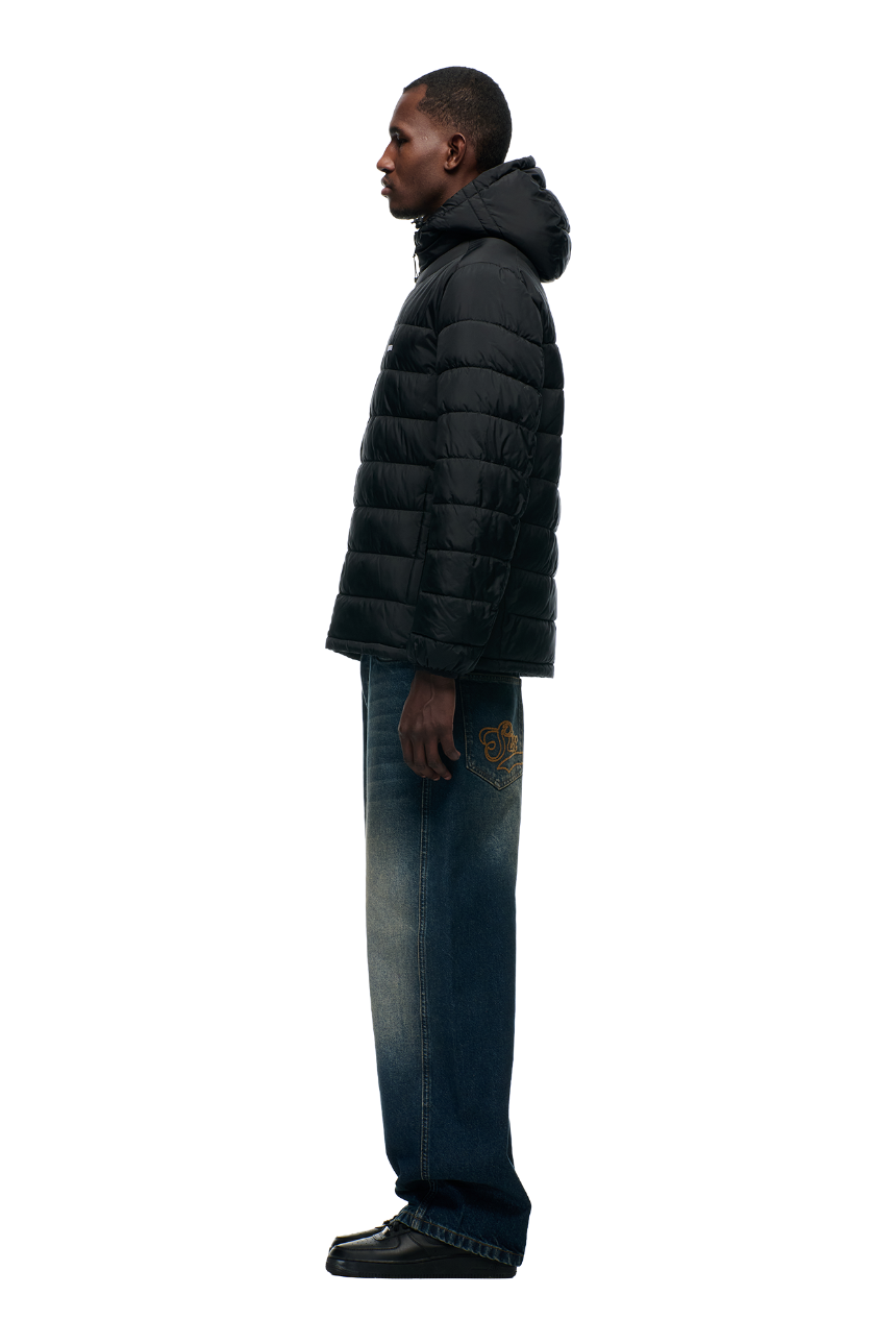A man stands in profile wearing the 6PM LIGHT WEIGHT PUFFER JACKET BLACK with a hood, wide-leg blue jeans with gold embroidery, and black shoes, set against a plain light background.