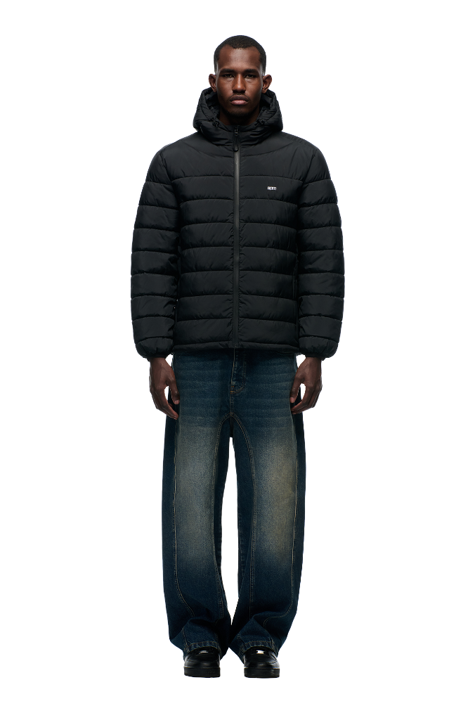 A man faces forward, wearing the 6PM LIGHT WEIGHT PUFFER JACKET BLACK with a hood, wide-leg blue jeans with faded areas, and black shoes, set against a plain light gray background.