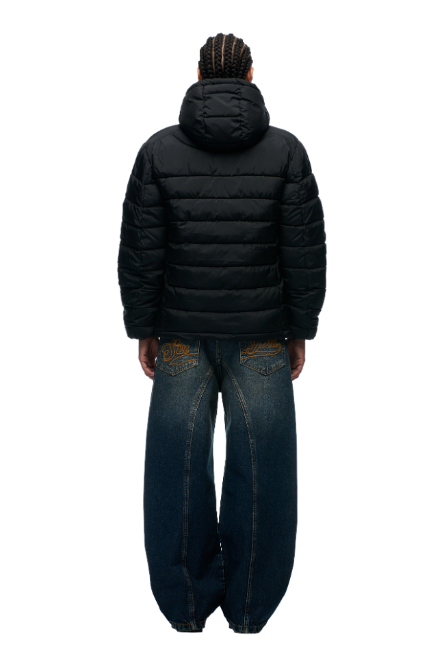A person with braided hair faces away, wearing the 6PM LIGHT WEIGHT PUFFER JACKET BLACK with a hood and loose-fitting blue jeans with detailed back pocket stitching, set against a plain white background.