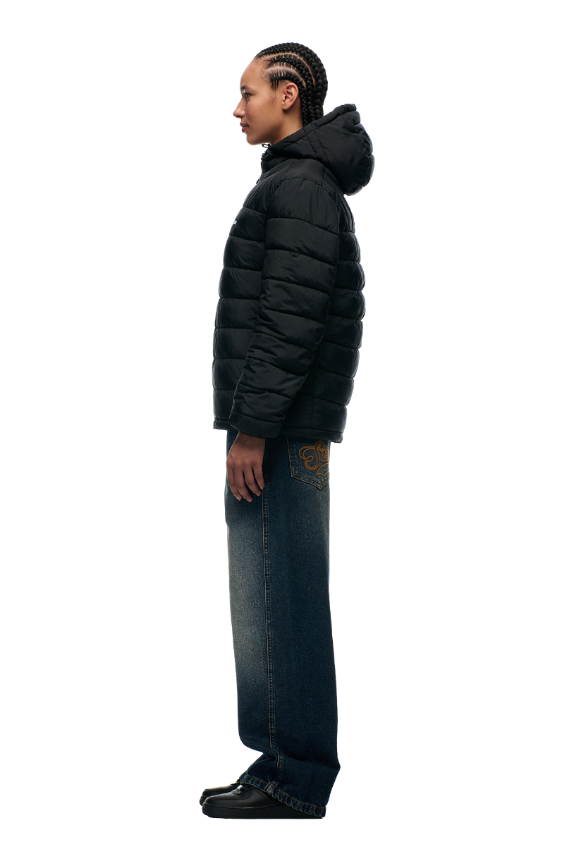A person in profile wears the 6PM LIGHT WEIGHT PUFFER JACKET BLACK with a hood, blue wide-leg jeans featuring embroidered pockets, and black shoes against a plain light background.