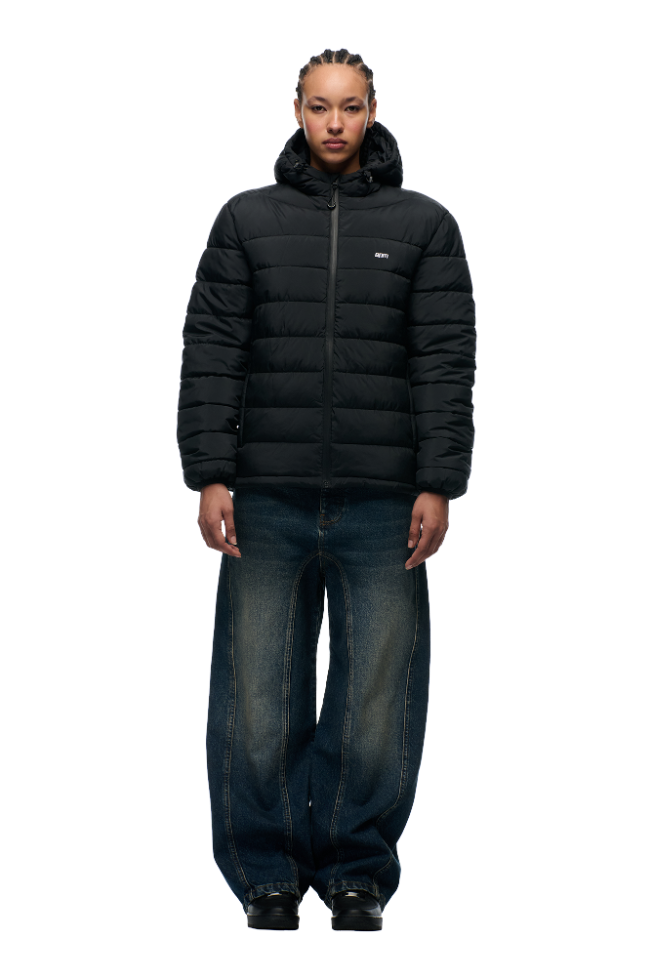 A person with braided hair wears the 6PM LIGHT WEIGHT PUFFER JACKET BLACK, wide-leg blue jeans, and black shoes, standing against a plain light gray background.