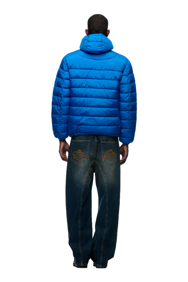 A person stands with their back to the camera, wearing the 6PM LIGHT WEIGHT PUFFER JACKET in Princess Blue, featuring an adjustable fit and hood, paired with baggy blue jeans with orange embroidery, against a plain light background.