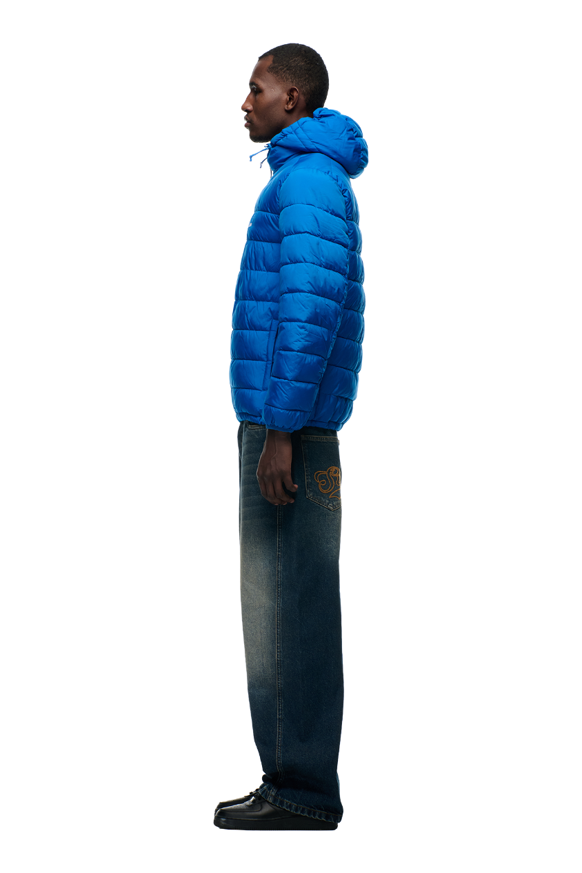 A person stands in profile against a white background, wearing the 6PM LIGHT WEIGHT PUFFER JACKET in Princess Blue, paired with loose blue jeans featuring a gold pocket design and black shoes.