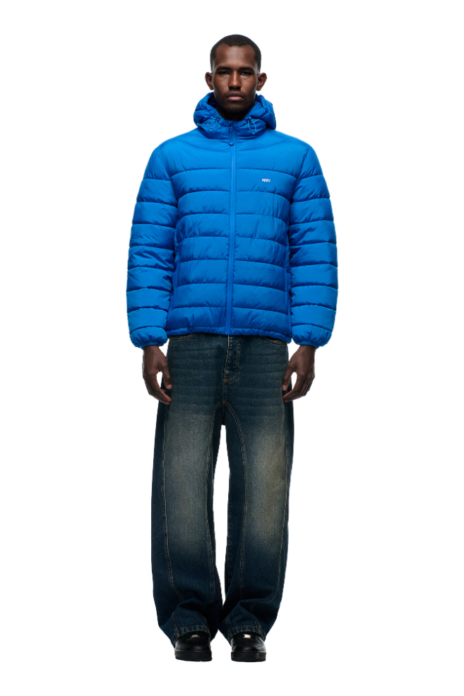 A man faces forward in a 6PM LIGHT WEIGHT PUFFER JACKET PRINCESS BLUE with a hood, wide-leg blue jeans, and black shoes against a plain white background.