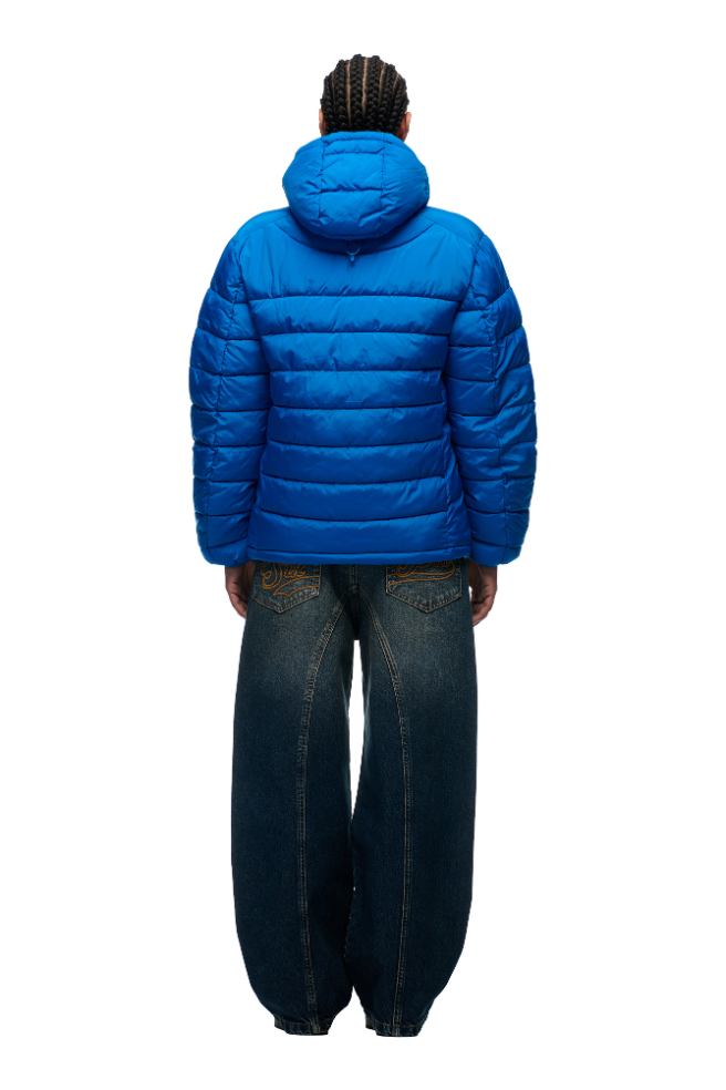 A person with braided hair wears the 6PM LIGHT WEIGHT PUFFER JACKET PRINCESS BLUE with a hood and loose, wide-leg blue jeans, standing with their back to the camera against a plain white background.
