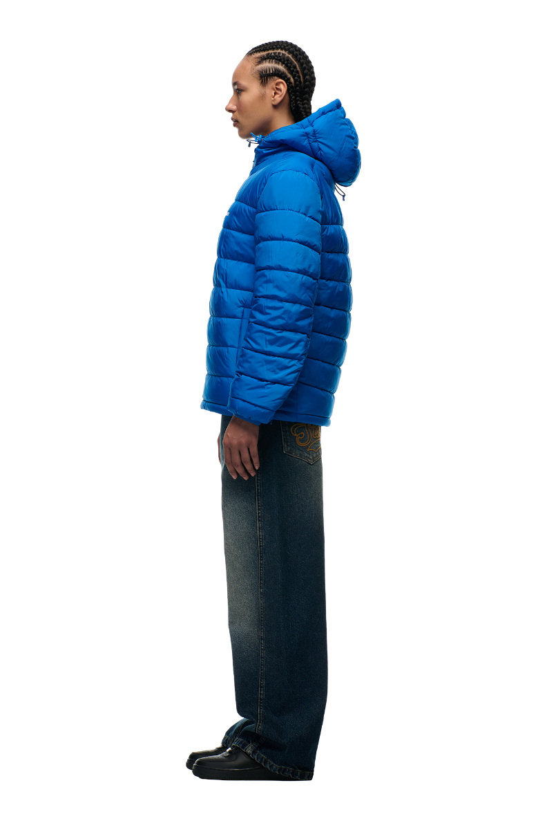 A person with braided hair stands in profile wearing the 6PM LIGHT WEIGHT PUFFER JACKET PRINCESS BLUE, wide-leg dark jeans, and black shoes against a plain white background.