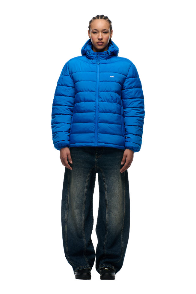A person faces forward wearing the 6PM LIGHT WEIGHT PUFFER JACKET PRINCESS BLUE with a hood and loose dark blue jeans against a plain white background.