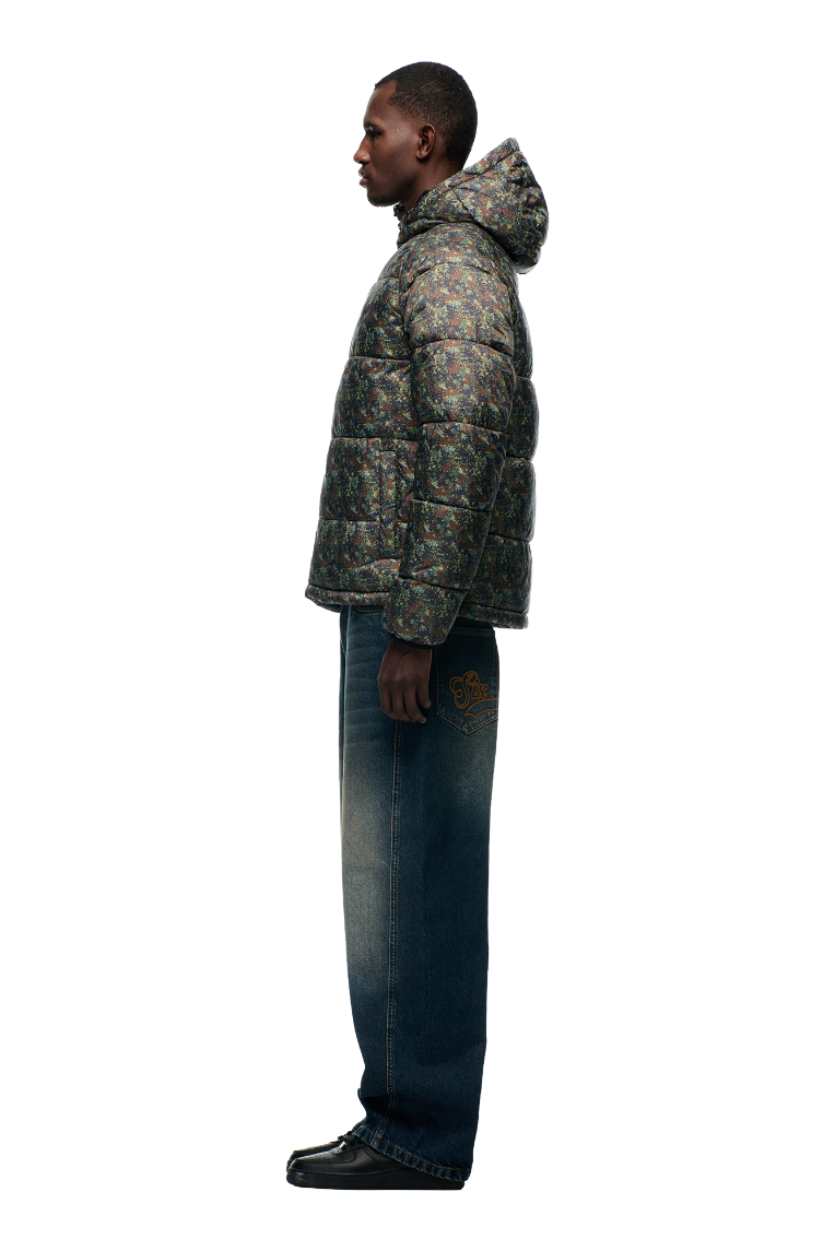 A man in profile wears the 6PM PUFFER JACKET MINI CAMO with an adjustable hood, loose blue jeans with an embroidered back pocket, and black shoes, all set against a plain white background.