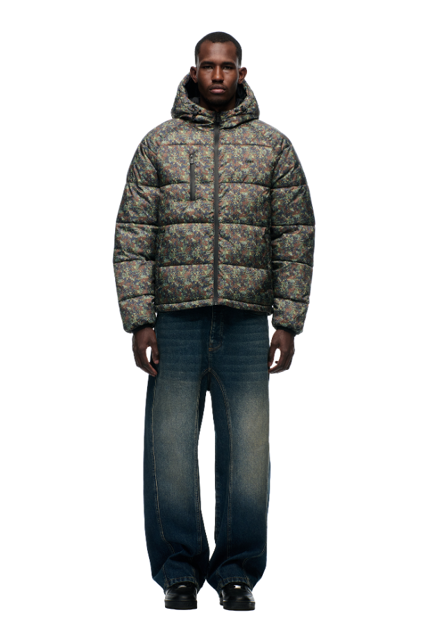 A man stands facing forward, dressed in a 6PM PUFFER JACKET MINI CAMO with an adjustable hood, wide-leg faded blue jeans, and black shoes against a plain white background.