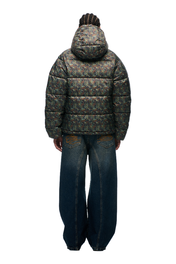 A person with braided hair stands with their back to the camera, wearing the 6PM PUFFER JACKET MINI CAMO—a water-repellent jacket with a multicolored floral pattern—and wide-leg blue jeans with embroidered back pockets.