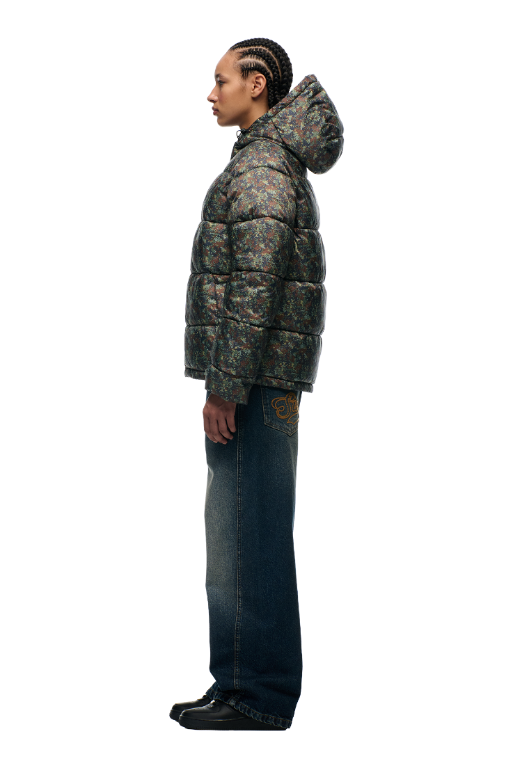 A person stands in profile wearing 6PM’s PUFFER JACKET MINI CAMO with an adjustable hood, wide-leg blue jeans with a yellow pocket graphic, and black shoes, all set against a plain white background.