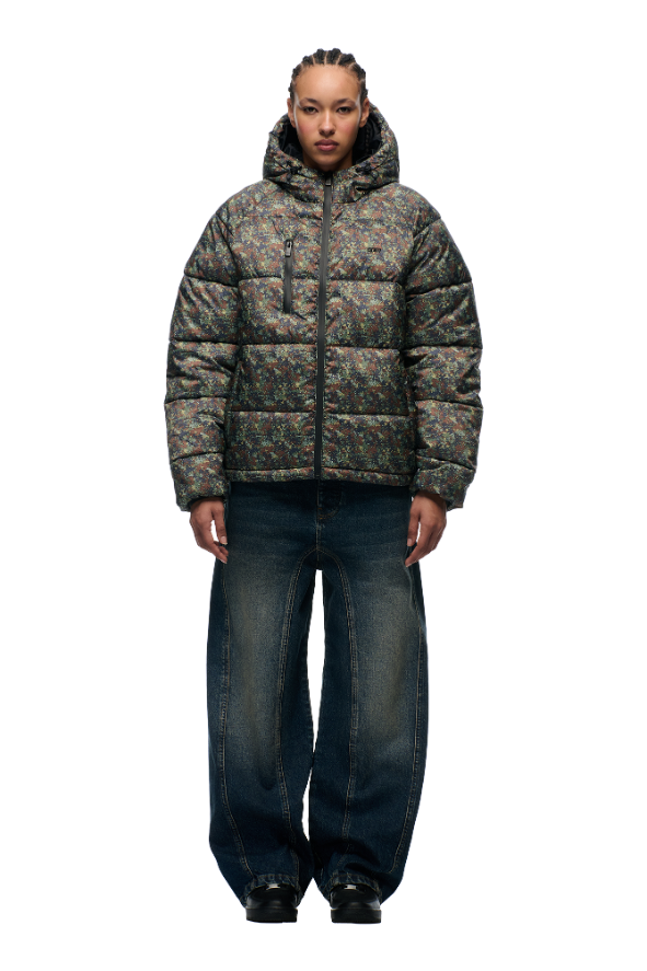 A person stands facing forward in the 6PM PUFFER JACKET MINI CAMO with an adjustable hood, paired with wide-leg blue jeans and black shoes, against a plain light background.