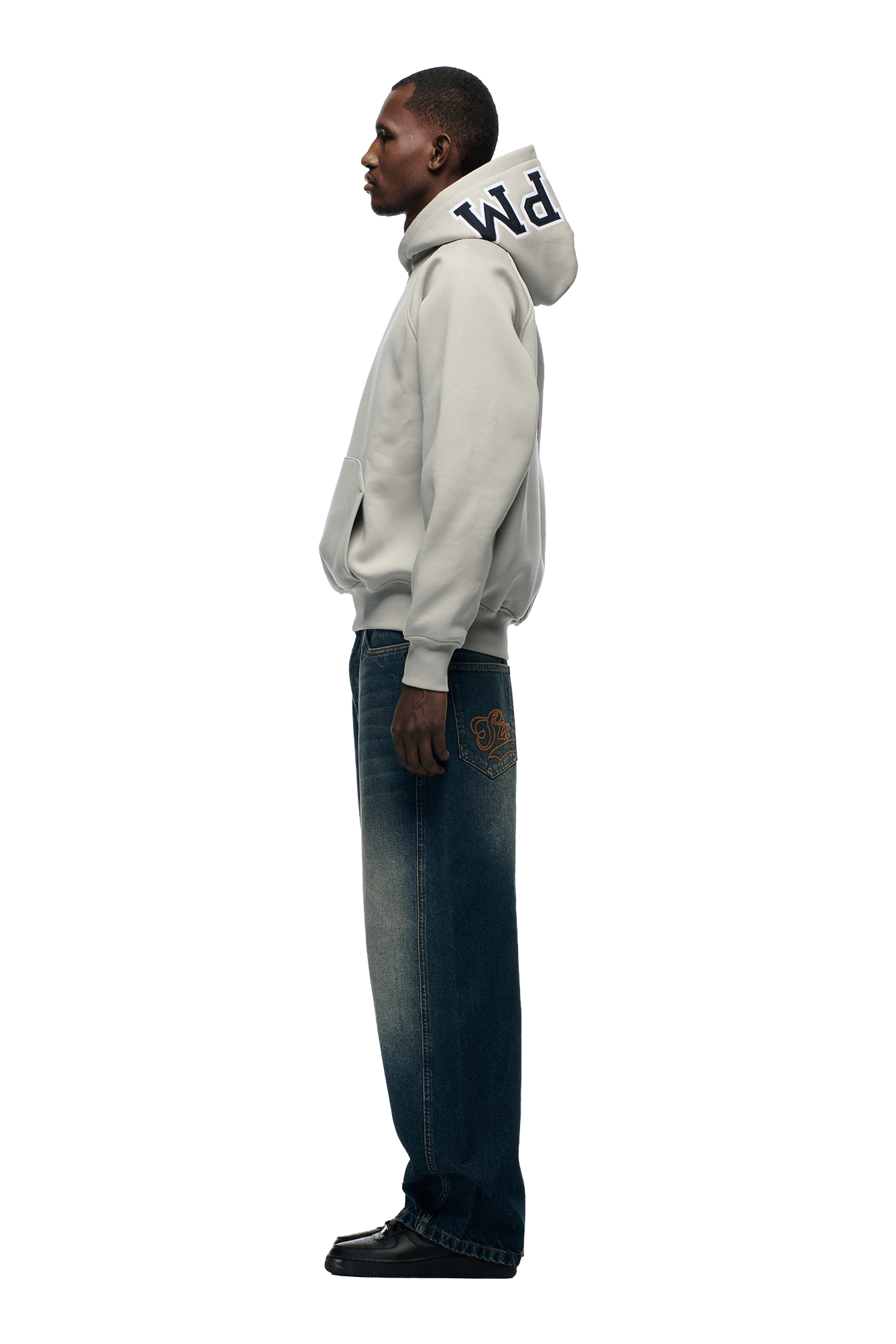 A man in profile against a white background wears the 6PM COLLEGE LETTER HOODIE in smoke grey, paired with blue jeans featuring a back pocket patch and black shoes.