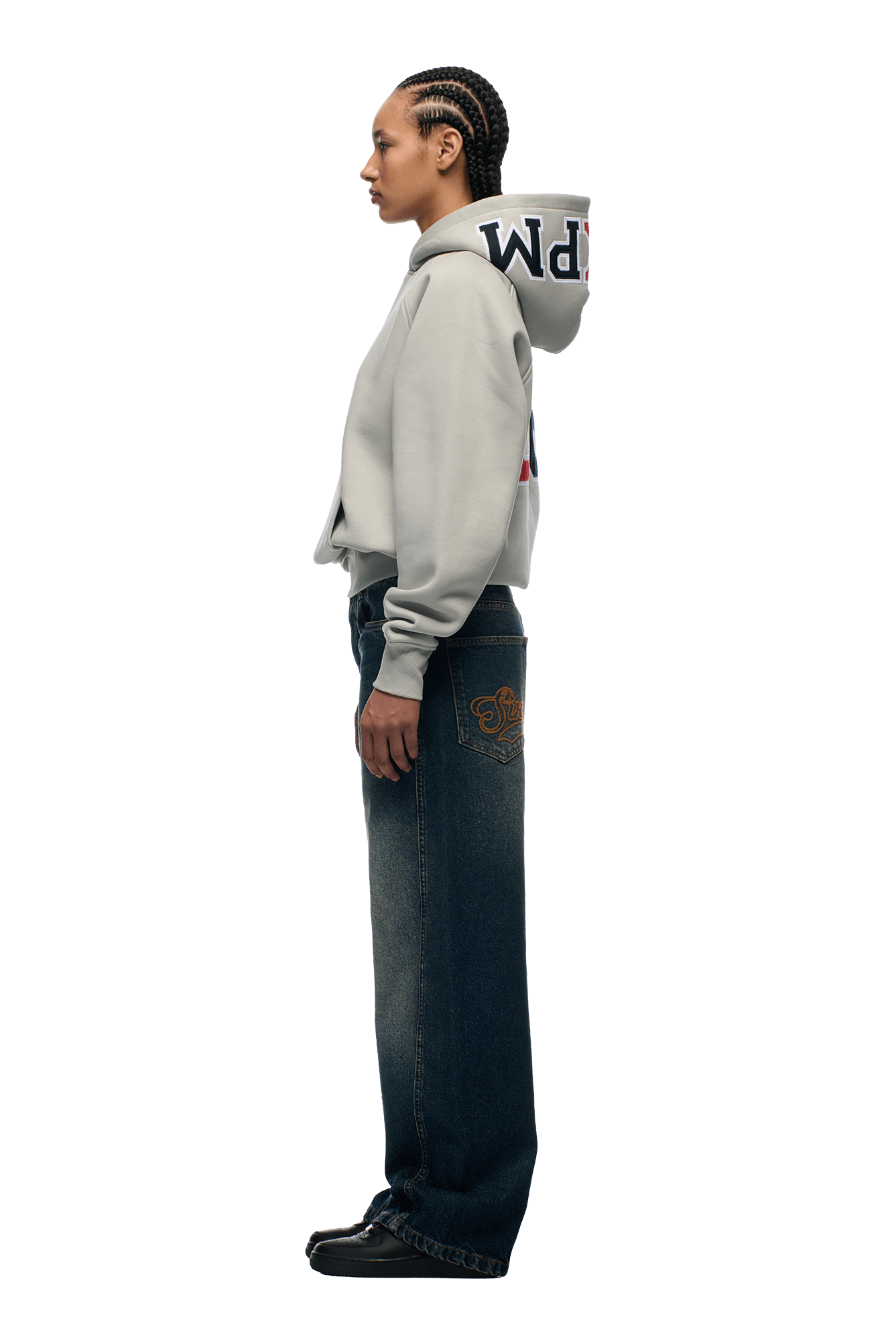 A person with braided hair stands in profile, wearing the 6PM COLLEGE LETTER HOODIE SMOKE GREY, wide-leg dark jeans featuring a gold pocket design, and black shoes, set against a plain white background.