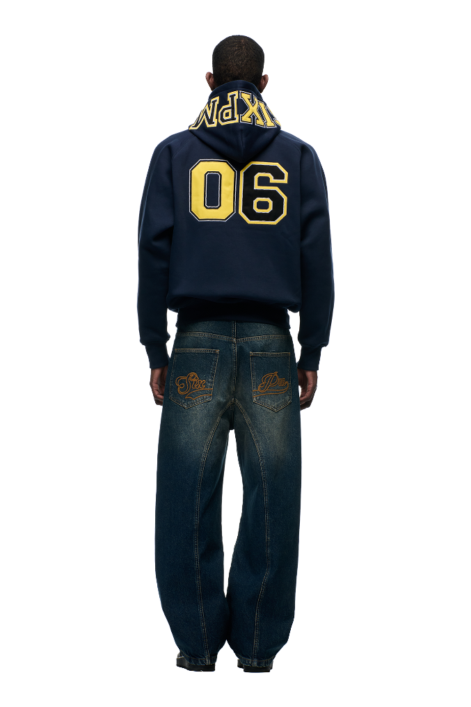 A person, seen from the back, wears the 6PM COLLEGE LETTER HOODIE MIDNIGHT BLUE with "90" and yellow 6PM lettering on the hood, paired with loose blue jeans featuring gold-embroidered back pockets, set against a white background.