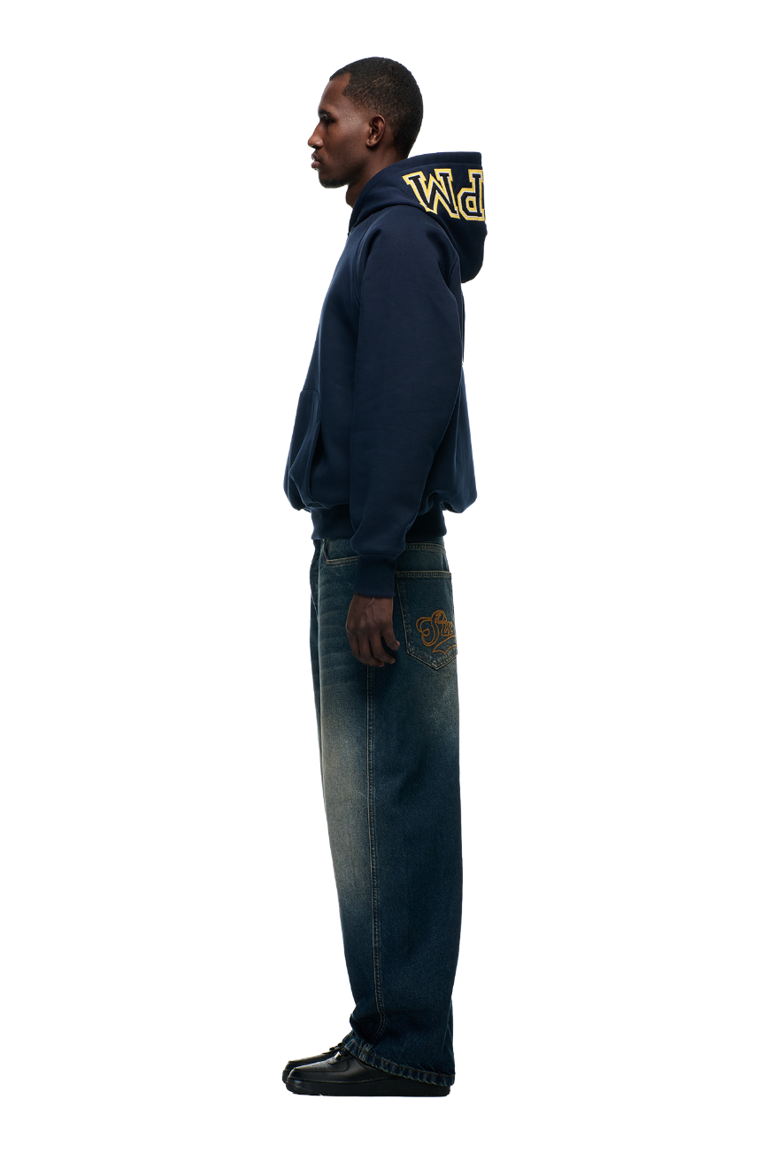A man in profile wears the 6PM COLLEGE LETTER HOODIE MIDNIGHT BLUE with yellow hood lettering, paired with loose blue jeans with back embroidery and black shoes, set against a plain white background.