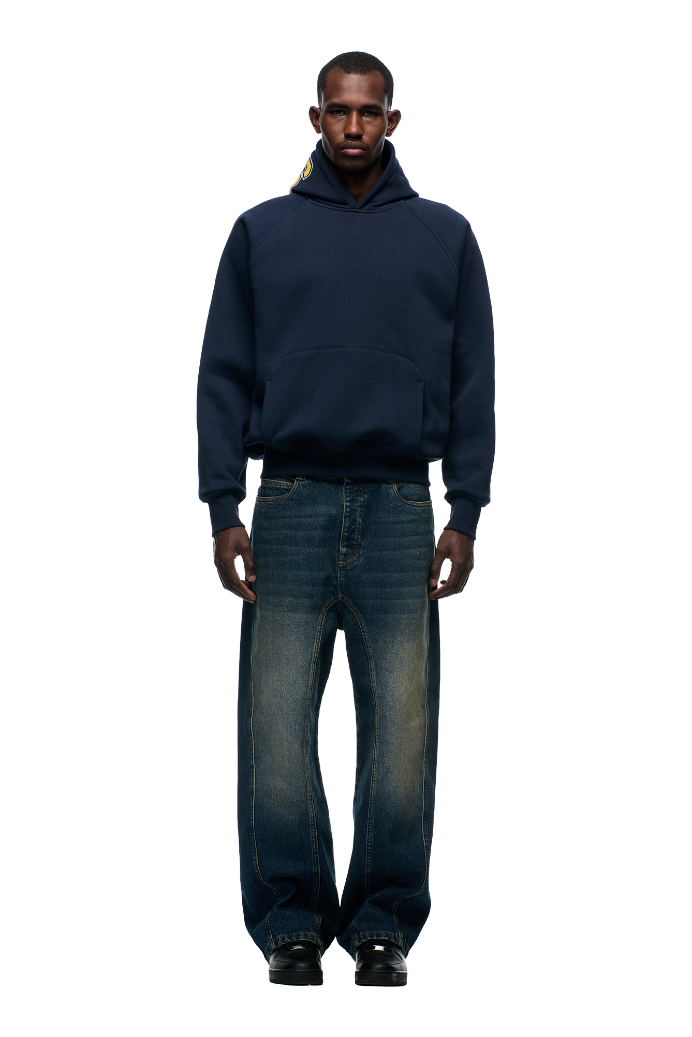 A man faces forward in a 6PM COLLEGE LETTER HOODIE MIDNIGHT BLUE, featuring back embroidery and 6PM lettering, paired with loose, faded blue jeans and black shoes, set against a plain light background.